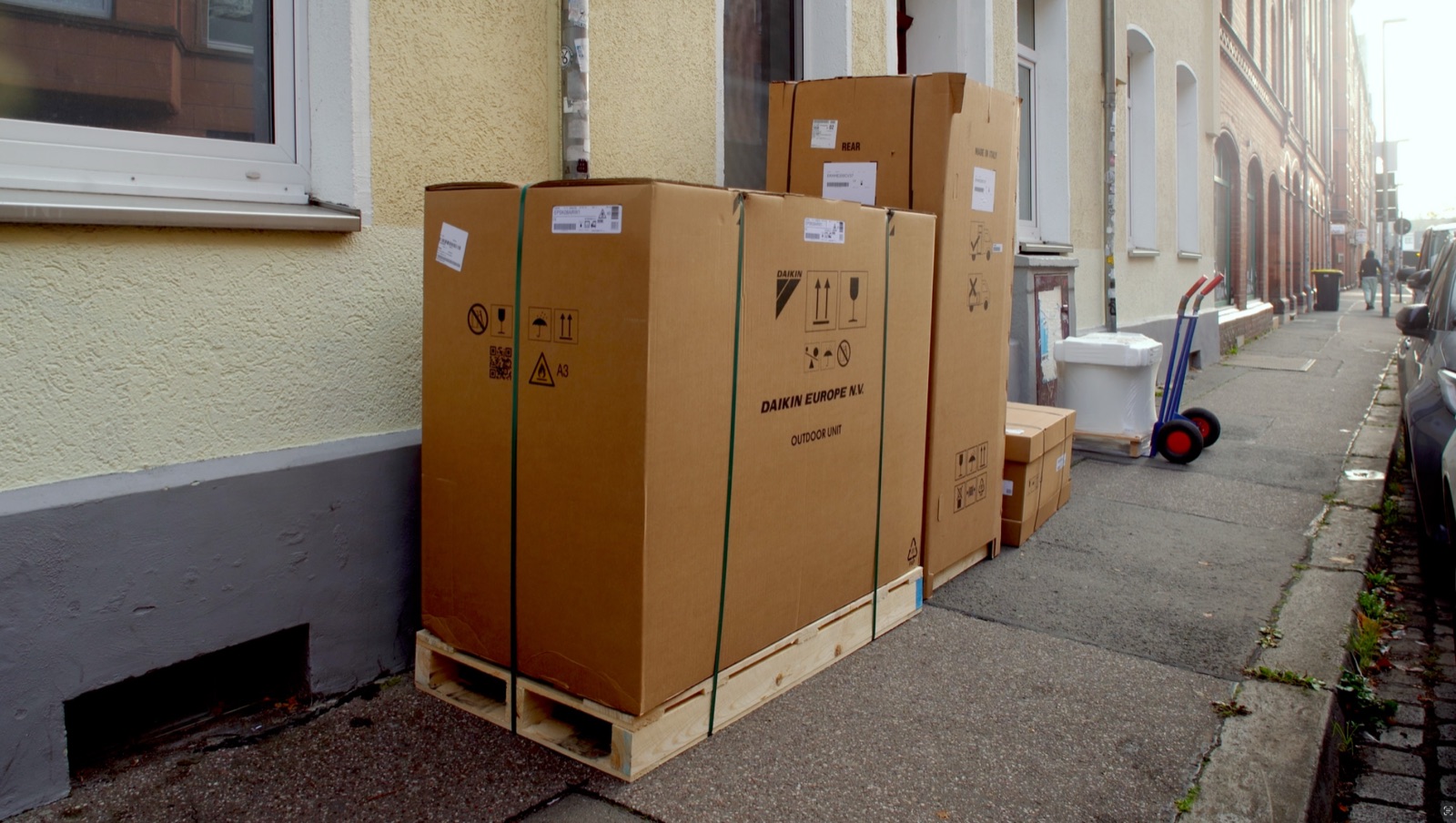 Verpackte Wärmepumpe und Anlagenteile auf Paletten vor einem Altbau, daneben eine Sackkarre